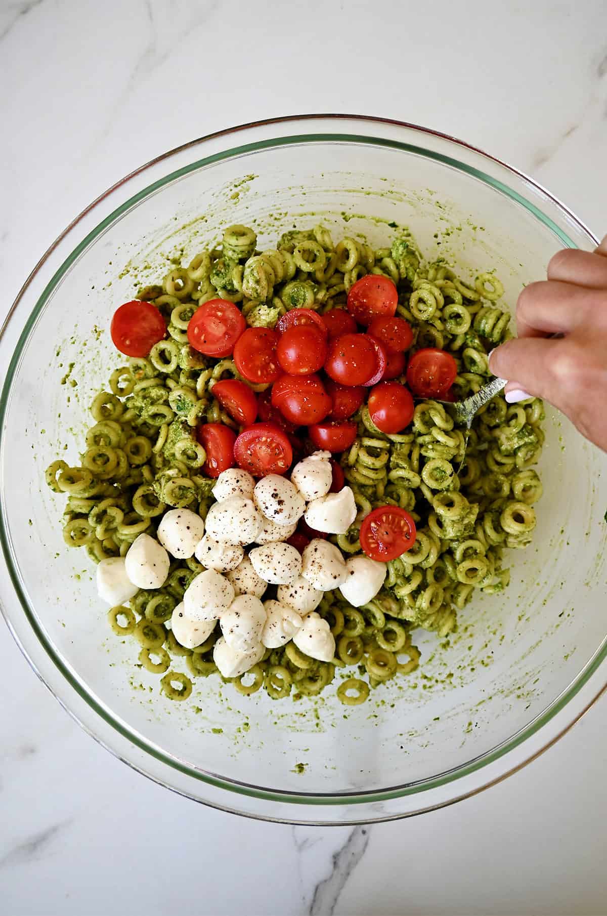 Halved cherry tomatoes and mini mozzarella balls atop pesto pasta in a large mixing bowl.