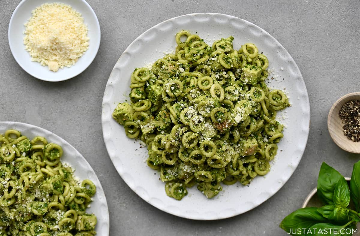 A white dinner plate containing pesto pasta with chicken next to a small plate with grated Parmesan cheese.