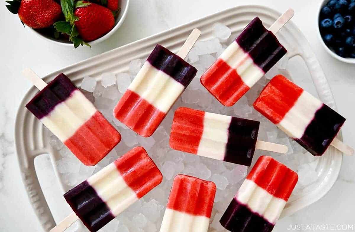 Red, white and blue popsicles over ice on a serving platter next to bowls containing fresh fruit.