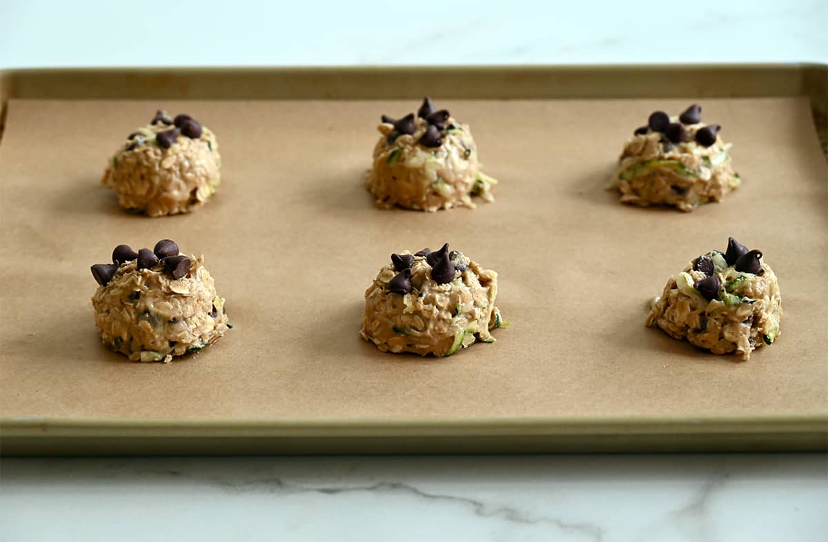 Unbaked mounds of zucchini oatmeal cookies topped with chocolate chips on a parchment paper-lined baking sheet.