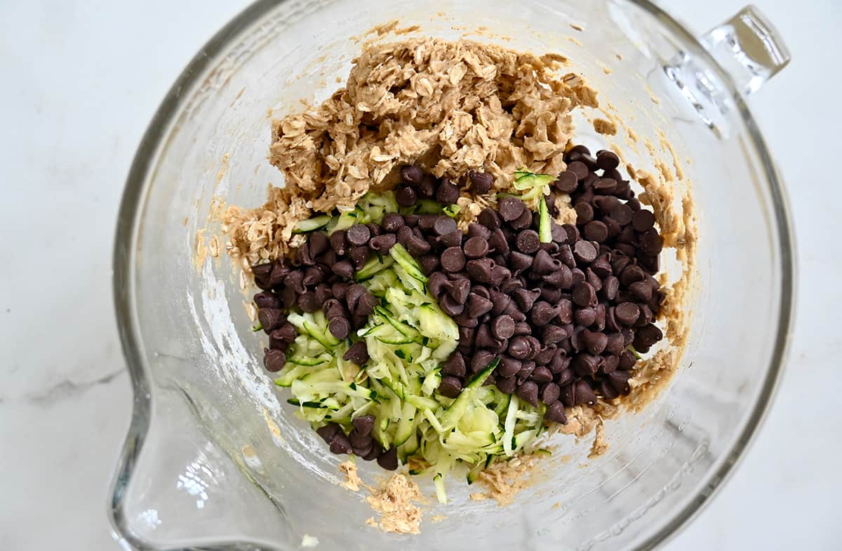 A top-down view of a large clear bowl of a stand mixer with oatmeal cookie batter topped with finely shredded zucchini and chocolate chips.