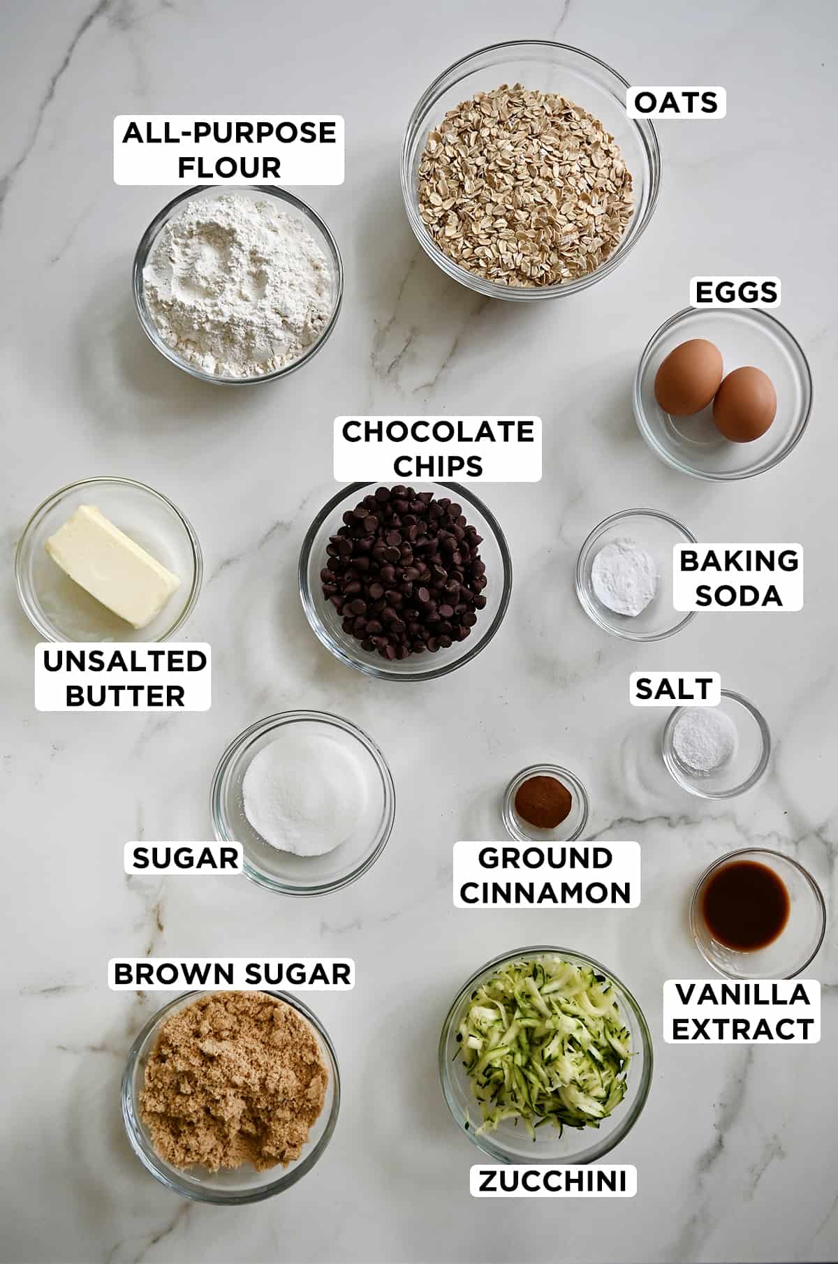 A top-down view of various sizes of clear bowls containing ingredients for zucchini oatmeal cookies, including all-purpose flour, oats, eggs, baking soda, salt, vanilla extract, ground cinnamon, shredded zucchini, brown sugar, sugar and unsalted butter. 