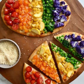 Two flatbread pizzas topped with a rainbow of veggies, including halved cherry tomatoes, diced orange bell pepper, fresh corn kernels, sliced summer squash, peas, broccoli florets, and purple cauliflower florets.