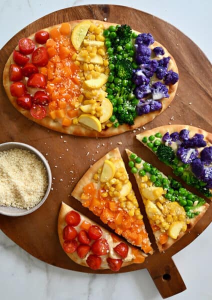 Two flatbread pizzas topped with a rainbow of veggies, including halved cherry tomatoes, diced orange bell pepper, fresh corn kernels, sliced summer squash, peas, broccoli florets, and purple cauliflower florets.