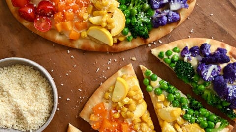 Two flatbread pizzas topped with a rainbow of veggies, including halved cherry tomatoes, diced orange bell pepper, fresh corn kernels, sliced summer squash, peas, broccoli florets, and purple cauliflower florets.