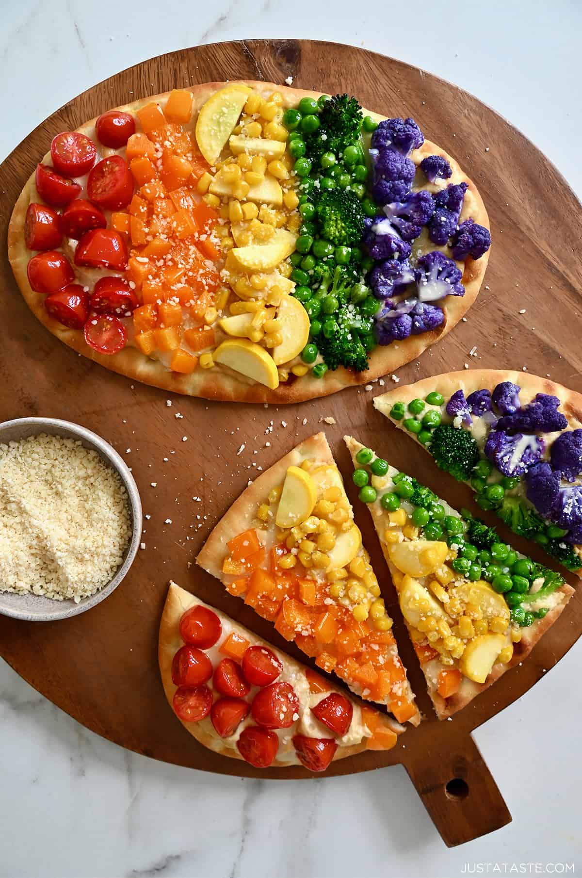 Two flatbread pizzas topped with a rainbow of veggies, including halved cherry tomatoes, diced orange bell pepper, fresh corn kernels, sliced summer squash, peas, broccoli florets, and purple cauliflower florets.