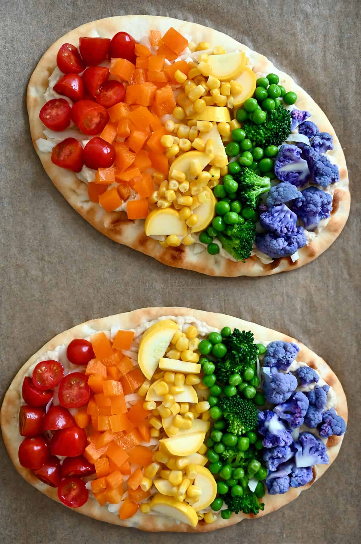 Two unbaked flatbread pizzas topped with halved cherry tomatoes, diced orange bell pepper, corn kernels, sliced summer squash, peas, broccoli florets and purple cauliflower florets. 