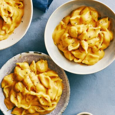 Two bowls containing creamy pumpkin macaroni and cheese next to a small bowl containing black pepper.