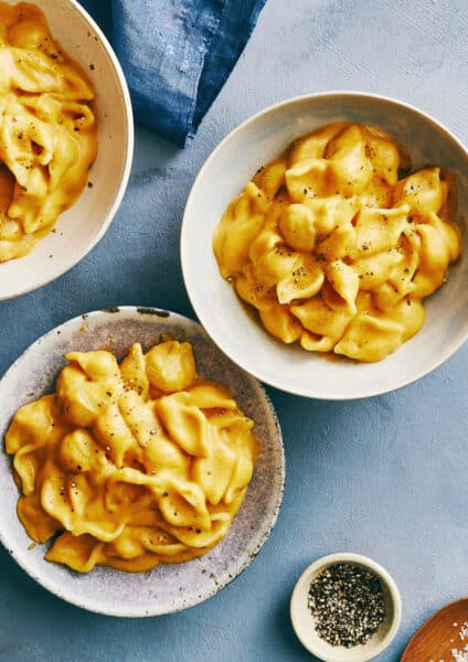 Two bowls containing creamy pumpkin macaroni and cheese next to a small bowl containing black pepper.