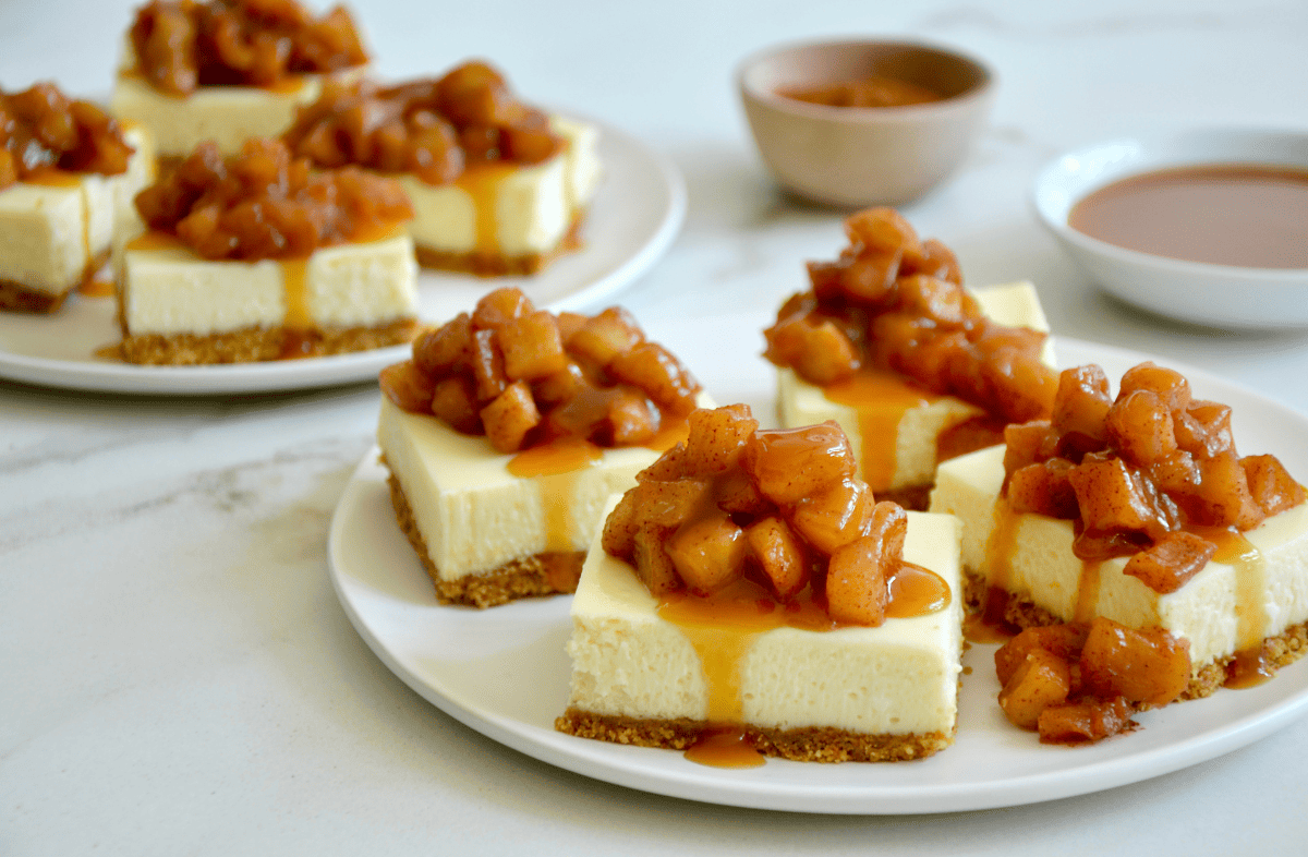 Two plates each with four cheesecake bars, topped with sauteed apples and caramel sauce.