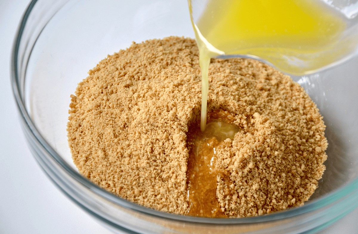 Melted butter is being poured over graham cracker crumbs in a clear glass bowl on a white background.