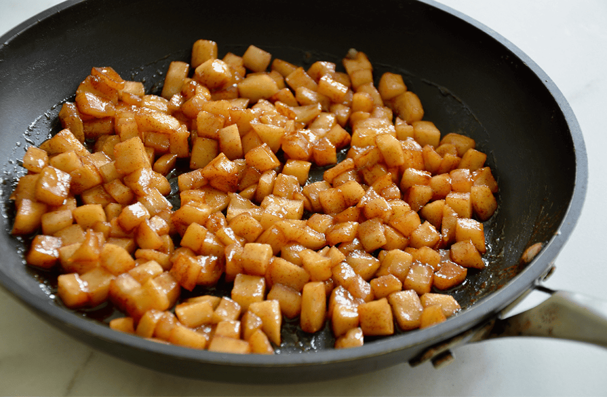 Diced apples that are cooked and caramelized are in a black skillet on a white countertop.