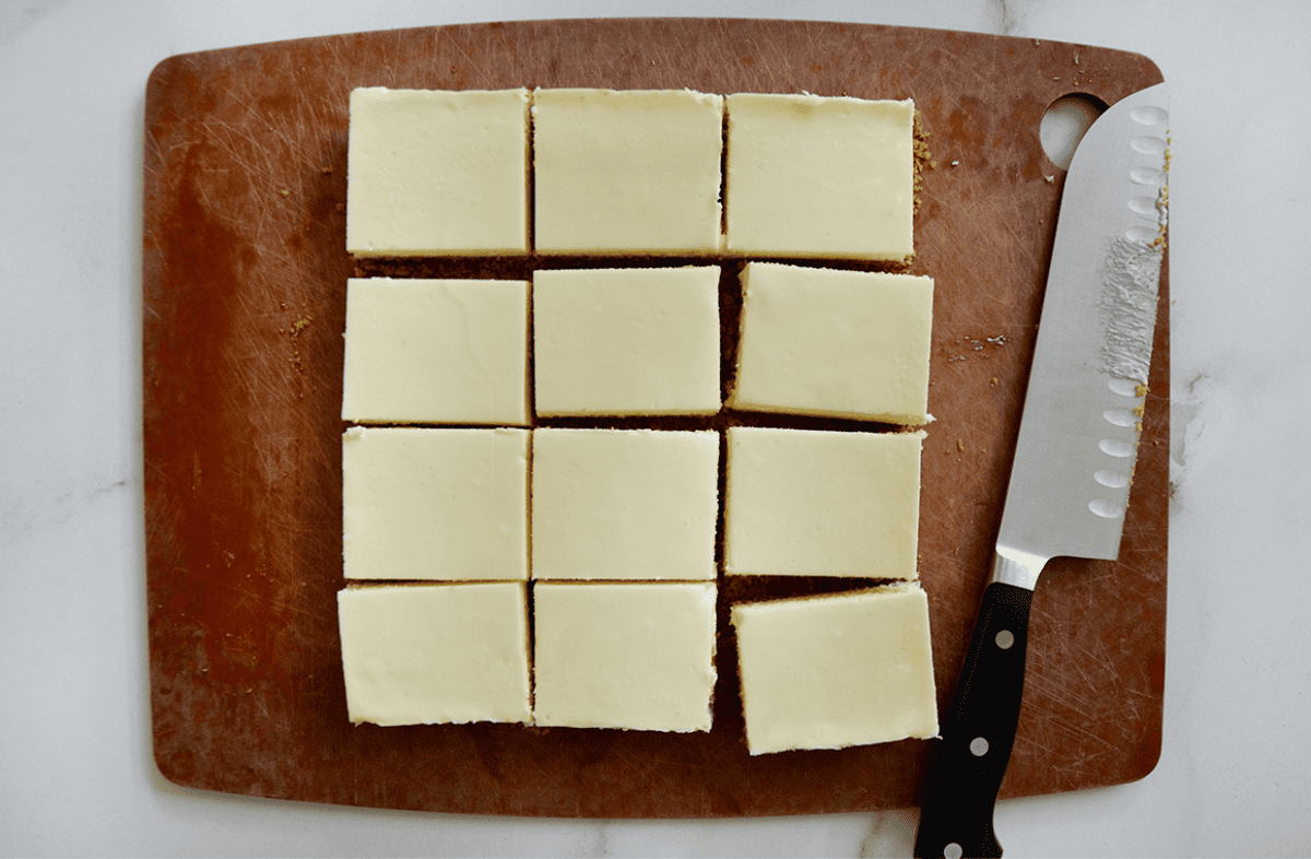 Cut cheesecake bars are on a brown rectangular cutting board on a white counter, with the santoku knife used to cut the bars next to them.