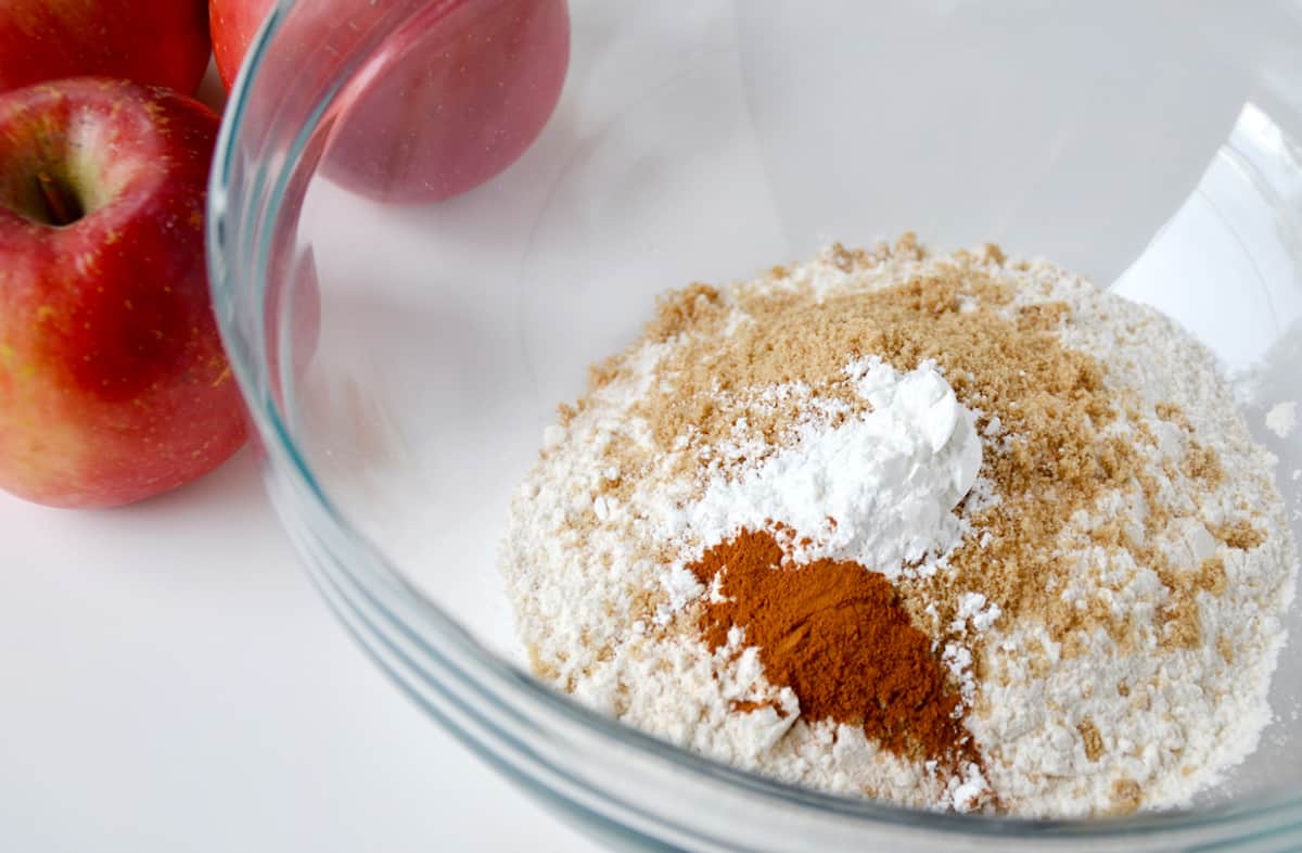 Dry ingredients (flour, cinnamon, brown sugar and baking powder) in a glass mixing bowl with two apples beside the bowl.