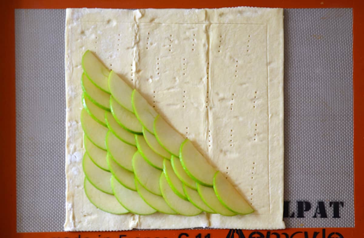 A square of puff pastry on a piece of Silpat with overlapping slices of apples covering half of the pastry.