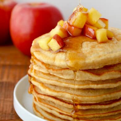 A stack of pancakes with sautéed apples on top and maple syrup being drizzled onto the pancakes.