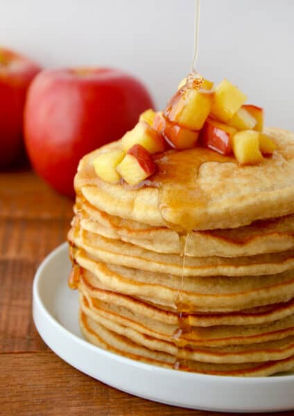 A stack of pancakes with sautéed apples on top and maple syrup being drizzled onto the pancakes.