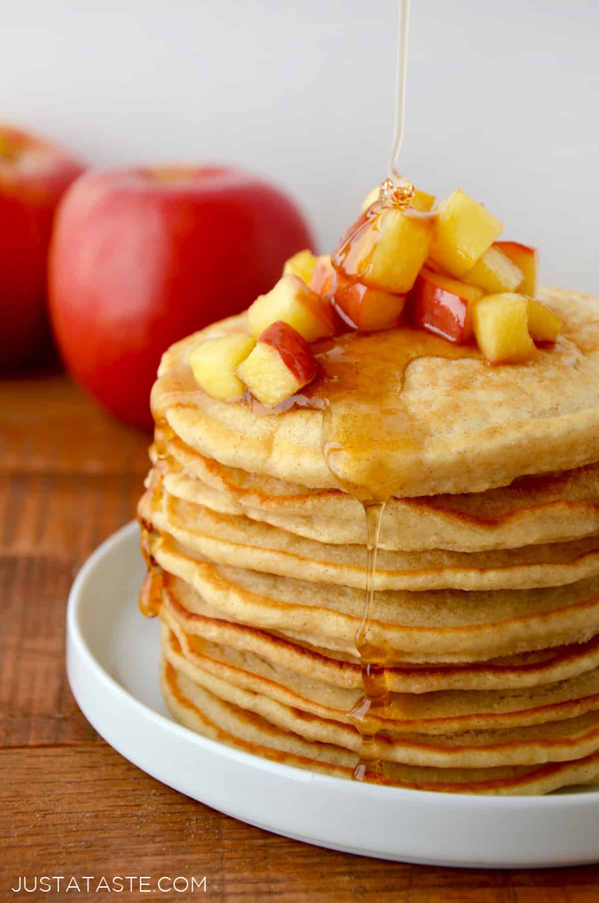 A stack of pancakes with sautéed apples on top and maple syrup being drizzled onto the pancakes.