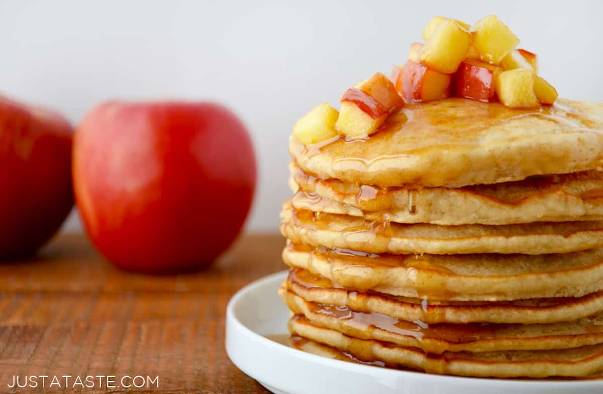 A plate of pancakes with sautéed apples and maple syrup on top and two apples behind it.