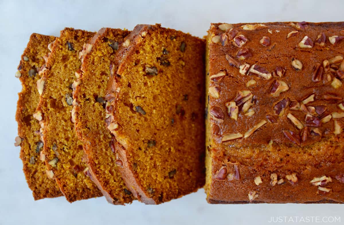 A loaf of pumpkin bread topped with chopped pecans and with four pieces sliced off of it and fanned out at the end of the loaf.