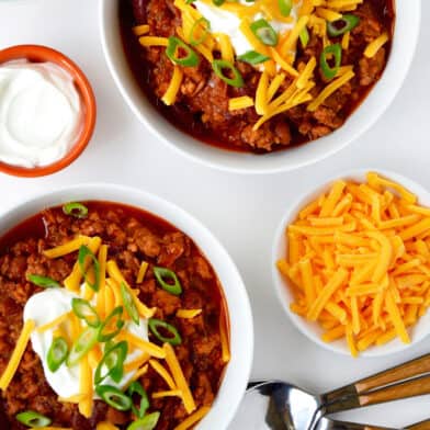 Two bowls of ground chicken chili topped with sour cream, grated cheese and sliced scallions. Spoons and small bowls of extra grated cheese, sour cream and sliced scallions surround the bowls of chili.