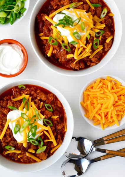 Two bowls of ground chicken chili topped with sour cream, grated cheese and sliced scallions. Spoons and small bowls of extra grated cheese, sour cream and sliced scallions surround the bowls of chili.