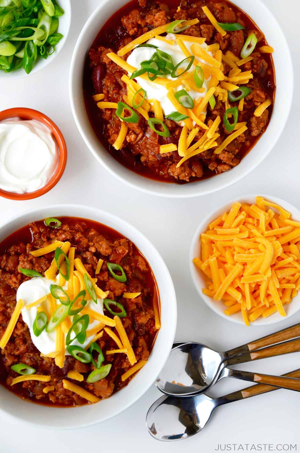 Two bowls of ground chicken chili topped with sour cream, grated cheese and sliced scallions. Spoons and small bowls of extra grated cheese, sour cream and sliced scallions surround the bowls of chili.