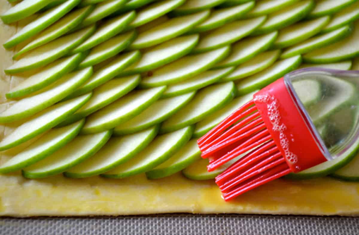 A pastry brush brushing the edges of an apple tart's puff pastry with egg wash.