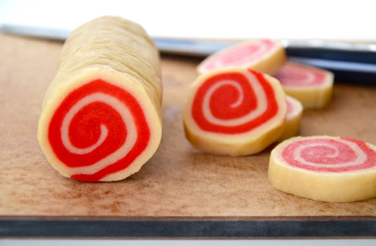 A log of red and white pinwheel cookie dough with five sliced cookies beside it.