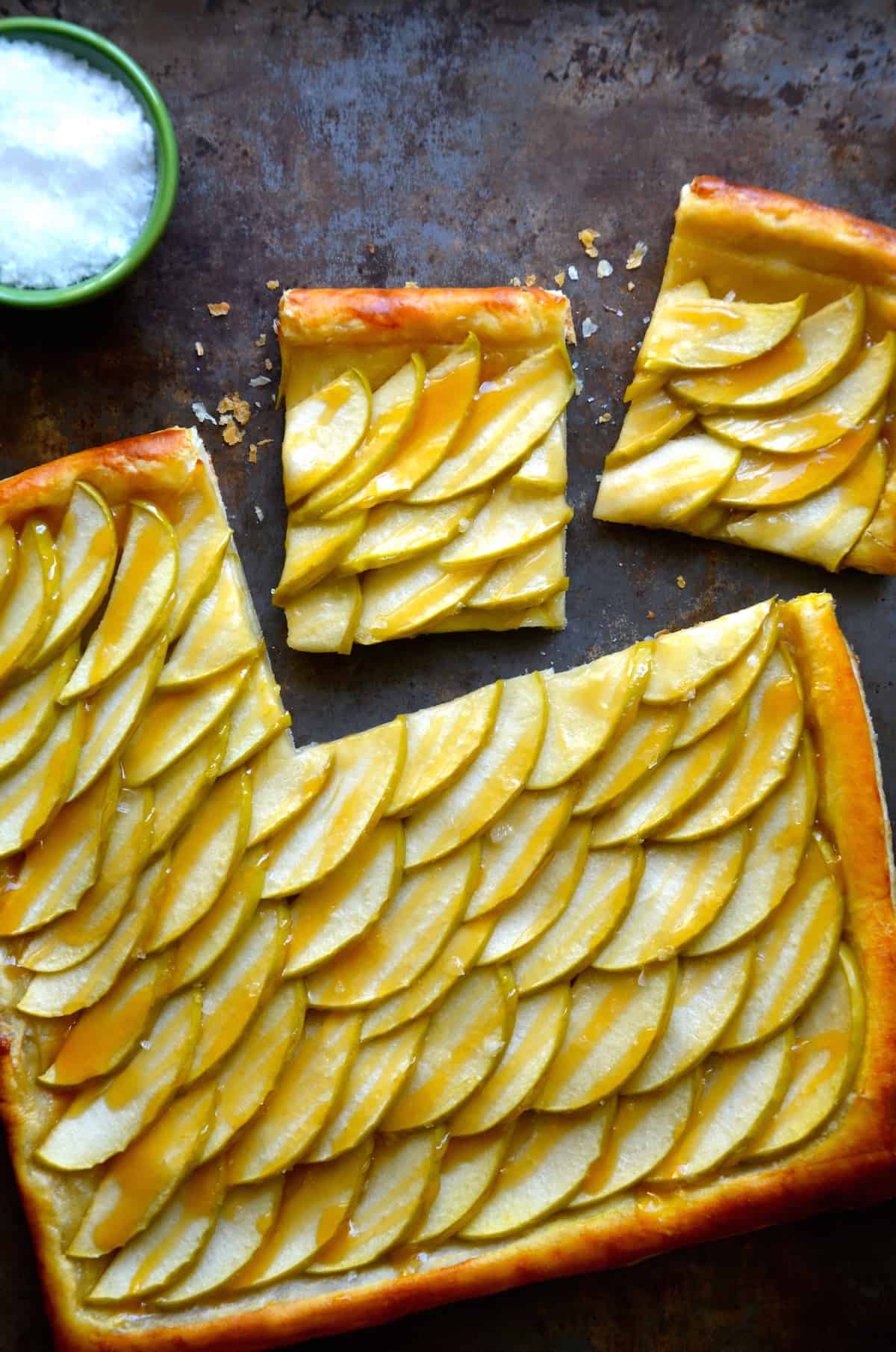 An apple tart made with puff pastry and drizzled with caramel sauce. There are two pieces sliced off of it and a small bowl of salt above the tart.