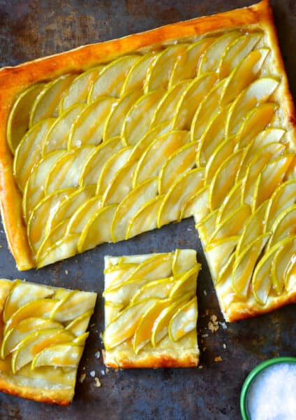 A salted caramel apple tart with two pieces sliced off of it and a small bowl of flakey salt next to it.