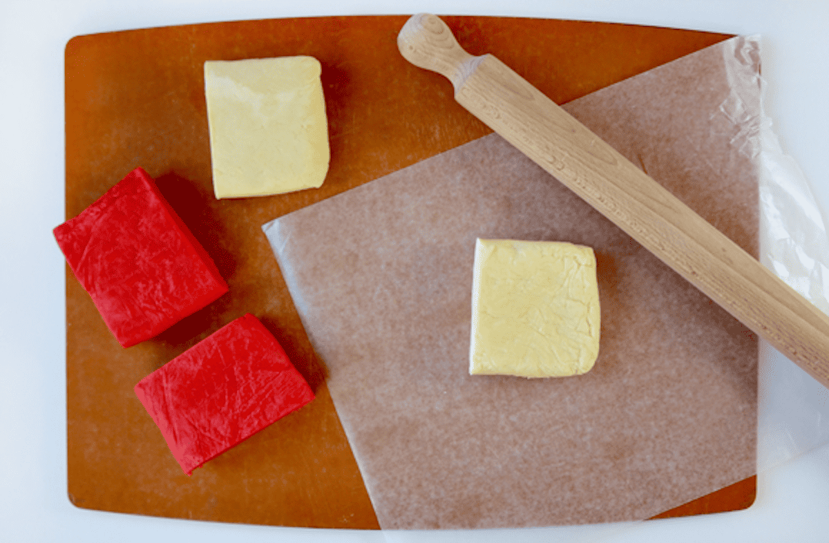 Four rectangles of dough (two white and two red) on a cutting board. One square of white dough is on a piece of wax paper, with a rolling pin sitting beside it.
