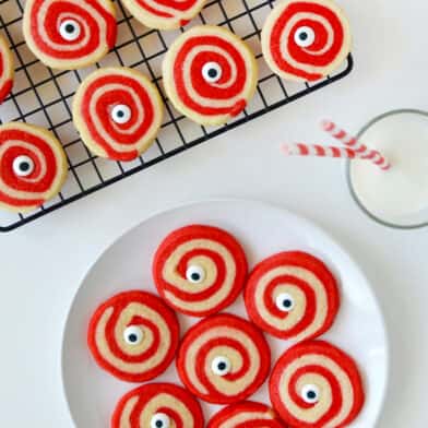 A plate of red and white Halloween pinwheel cookies with monster eyes and a glass of milk and a cooling rack with more cookies on it above it.