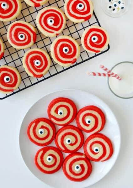 A plate of red and white Halloween pinwheel cookies with monster eyes and a glass of milk and a cooling rack with more cookies on it above it.