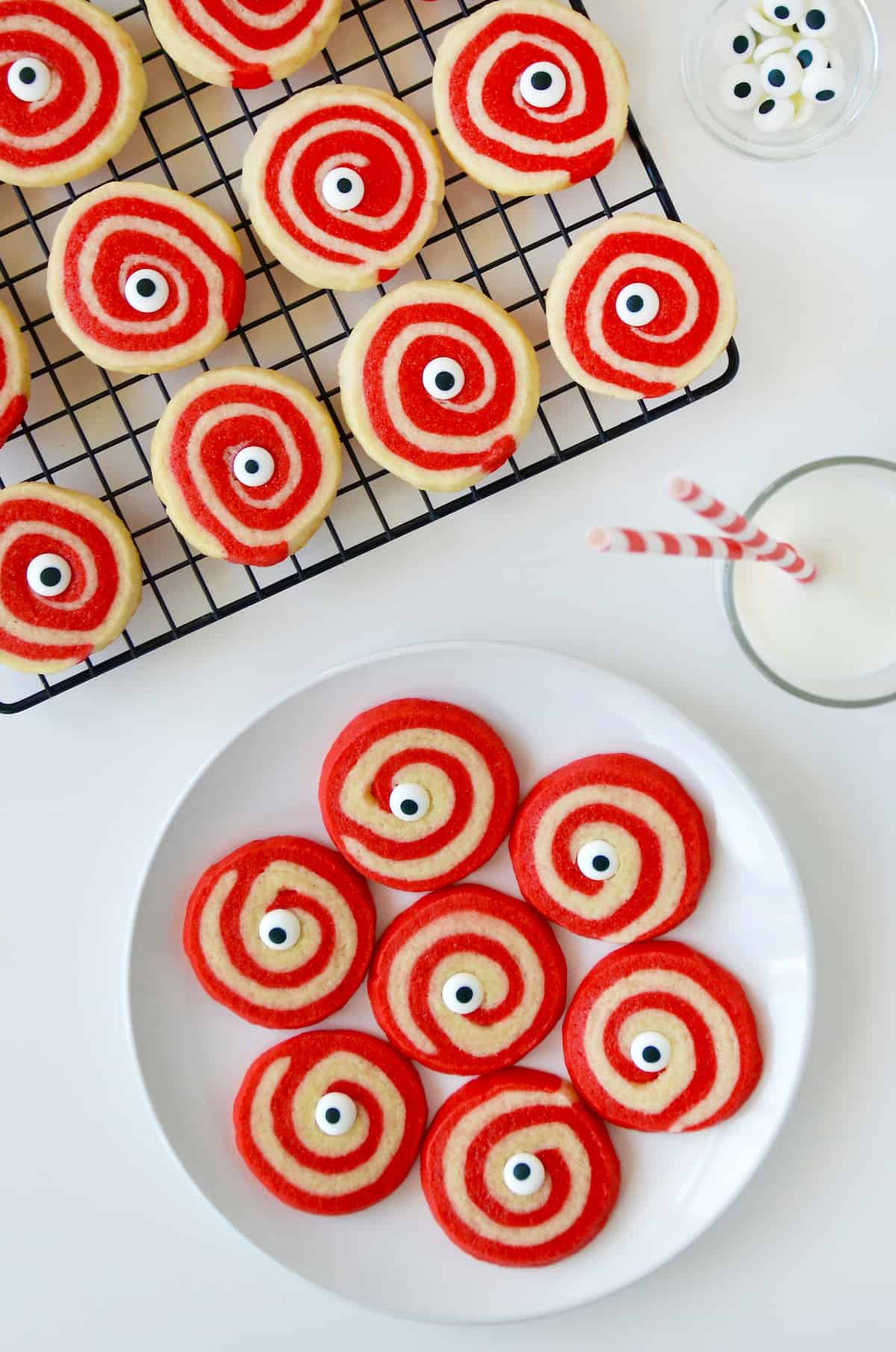 A plate of red and white Halloween pinwheel cookies with monster eyes and a glass of milk and a cooling rack with more cookies on it above it.