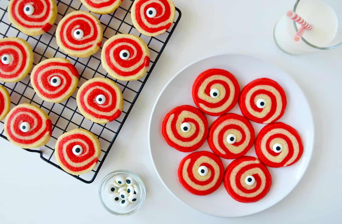 A plate of Halloween sugar cookies with a small bowl of candy eyeballs, a glass of milk and a cooling rack with more cookies surrounding it.