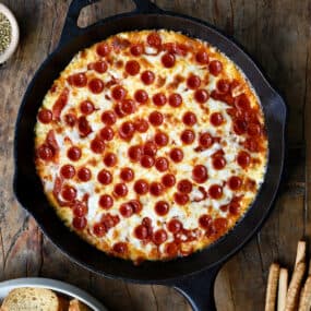 Pepperoni pizza dip in a cast iron skillet next to a small bowl containing crispy baguette toasts.