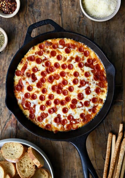 Pepperoni pizza dip in a cast iron skillet next to a small bowl containing crispy baguette toasts.