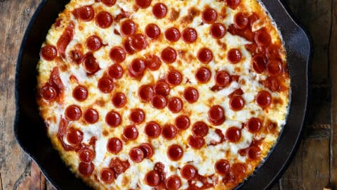 Pepperoni pizza dip in a cast iron skillet next to a small bowl containing crispy baguette toasts.