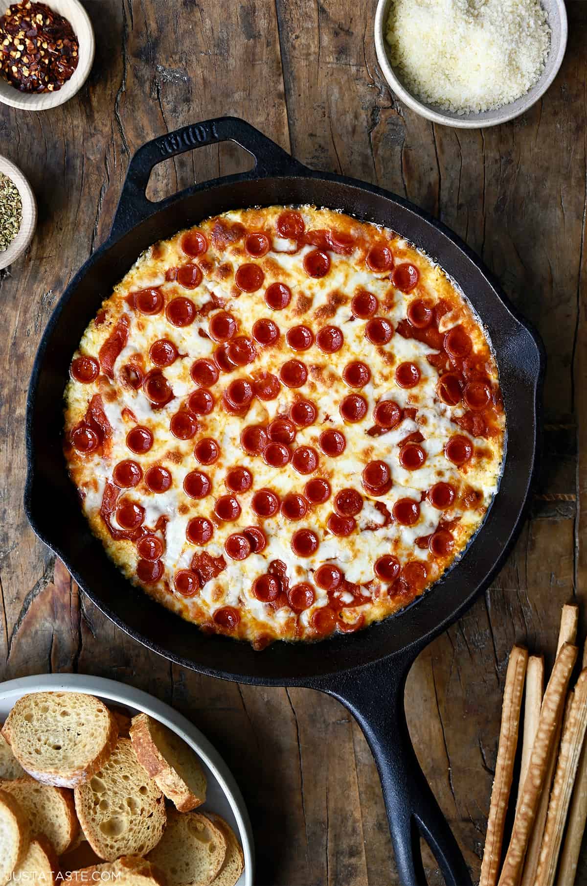 Pepperoni pizza dip in a cast iron skillet next to a small bowl containing crispy baguette toasts.