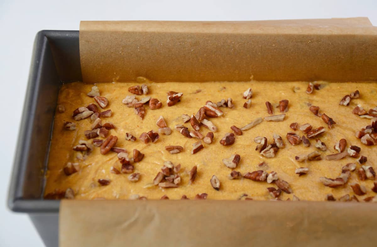 A loaf pan lined with parchment paper containing pumpkin bread batter topped with chopped pecans. 