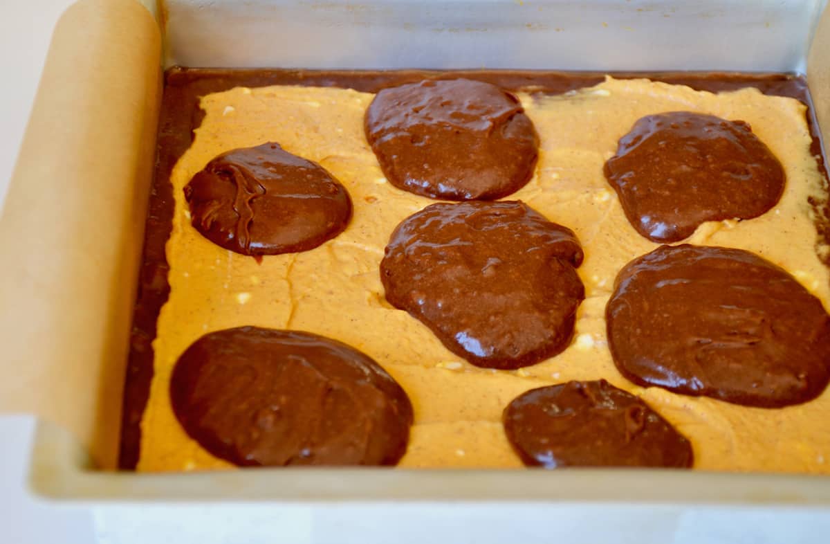 A pan of pumpkin cheesecake brownies with a layer of brownie batter, a layer of pumpkin cheesecake and dollops of brownie batter on top.