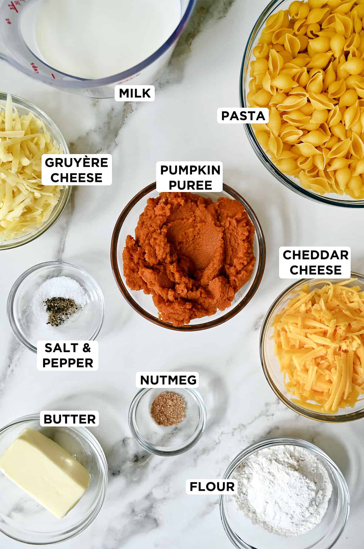 Various sizes of clear bowls containing milk, pasta shells, pumpkin puree, cheddar cheese, nutmeg, salt and pepper, butter, flour and gruyere cheese.