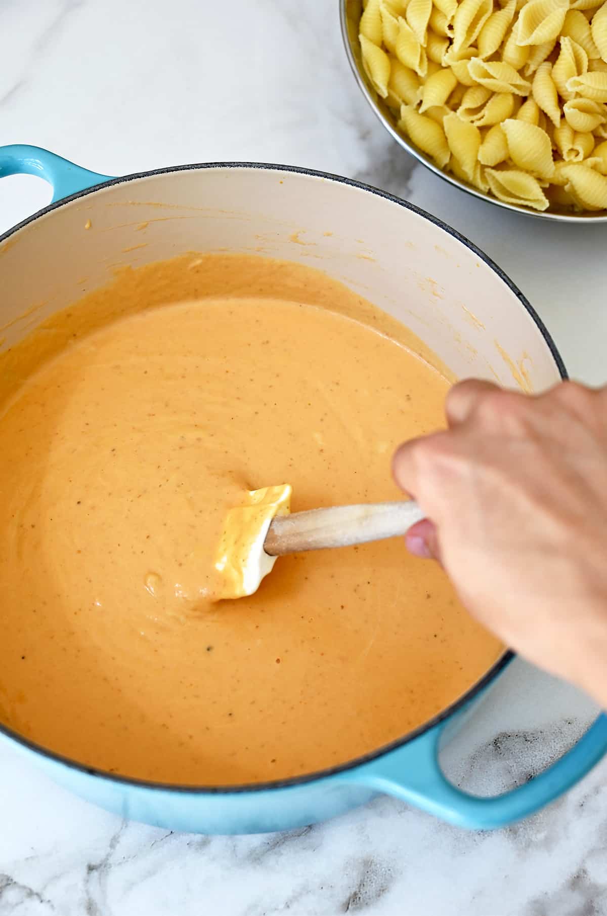 A hand holds a spatula into a large stockpot containing pumpkin cheese sauce.