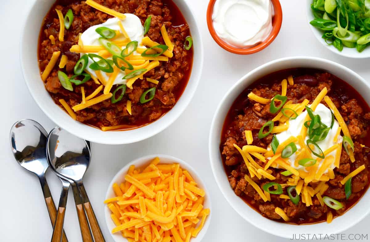Two bowls of chili topped with grated cheese, sliced scallions and sour cream. Spoons and bowls of grated cheese, sour cream and sliced scallions are next to the chili.