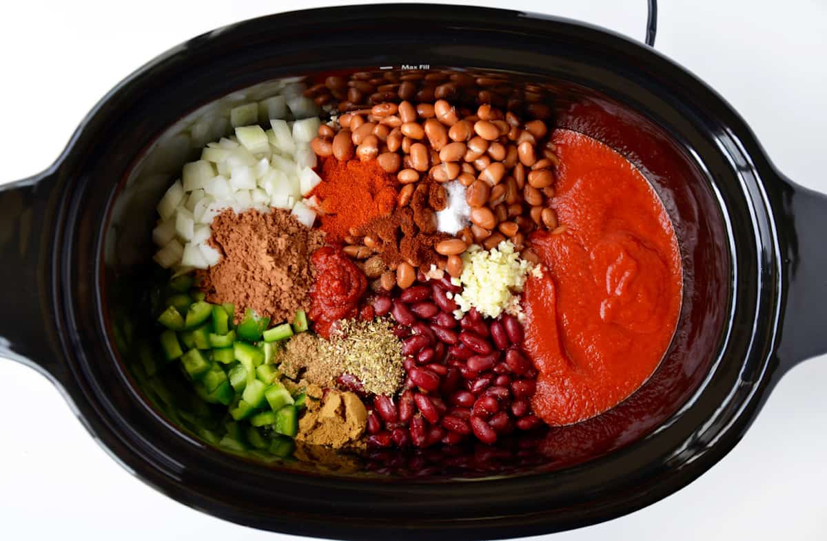 A slow cooker with tomato sauce and paste, beans, minced garlic, spices, chopped onions and chopped bell pepper inside of it.