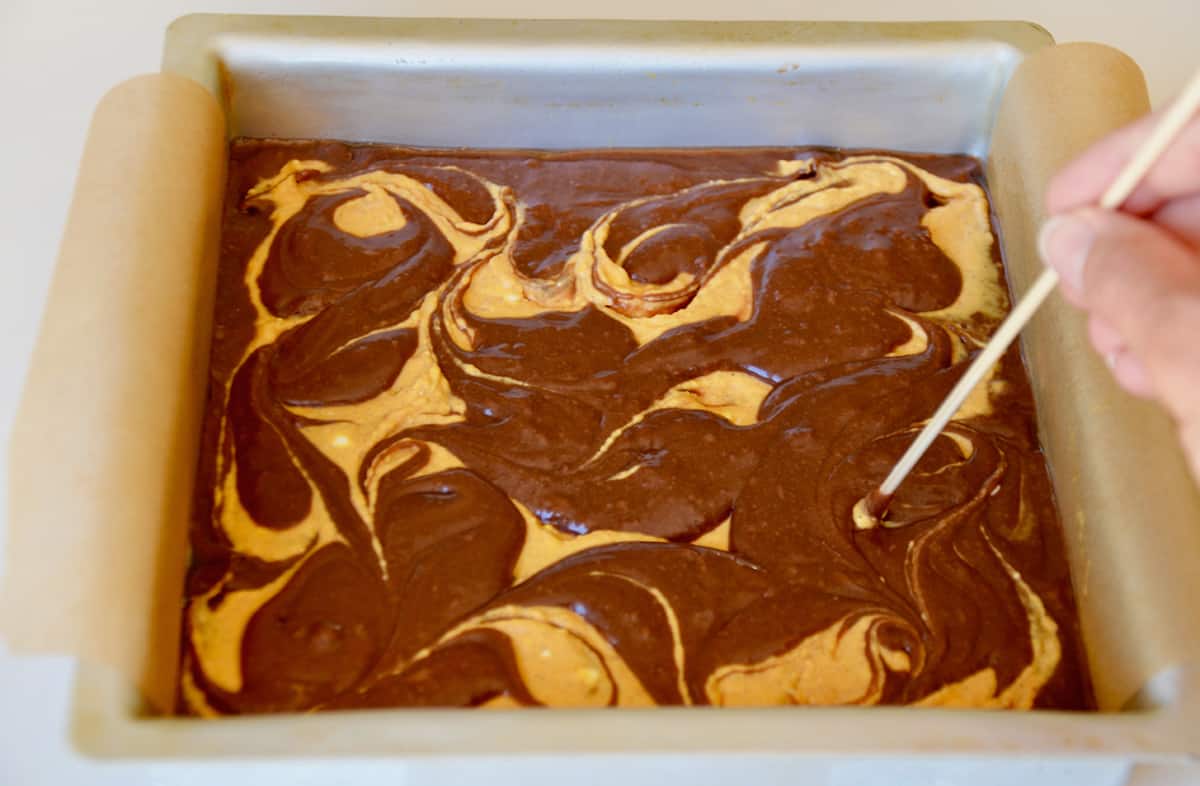 A person using a wooden skewer to swirl brownie and pumpkin cheesecake batter together.