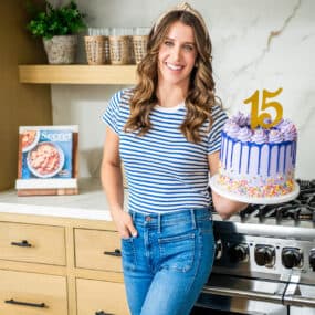 Chef Kelly Senyei standing in her kitchen holding a purple cake with the number "15" on it