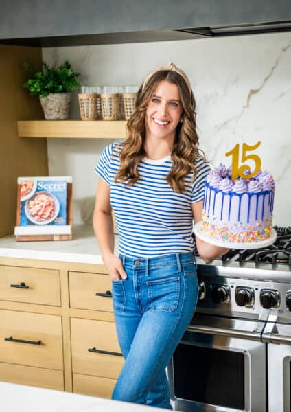 Chef Kelly Senyei standing in her kitchen holding a purple cake with the number "15" on it
