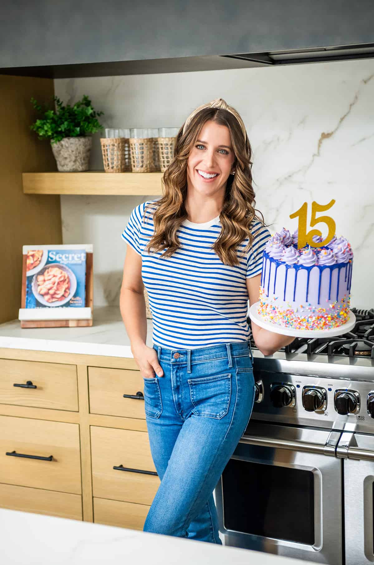 Chef Kelly Senyei standing in her kitchen holding a purple cake with the number "15" on it.